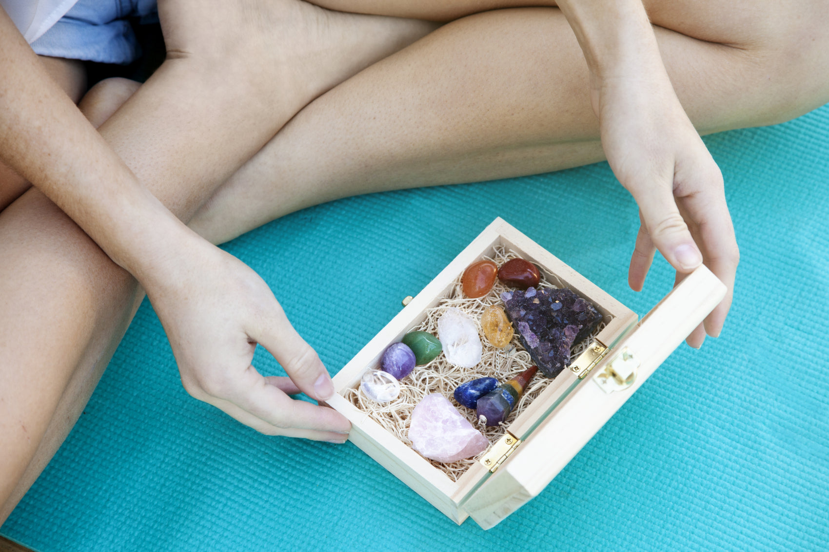 Person sitting on a yoga mat holding a wooden box filled with various healing crystals.