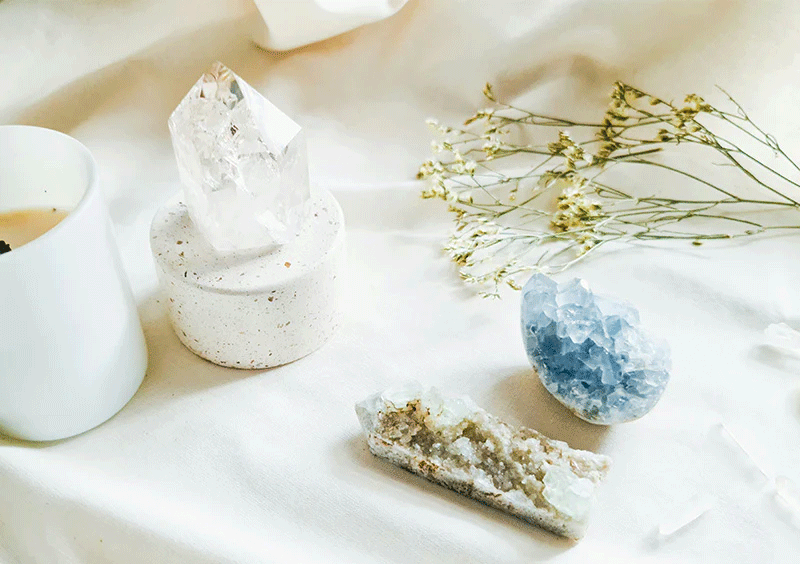 A clear quartz crystal and celestite stone placed on a white cloth with a candle and dried flowers
