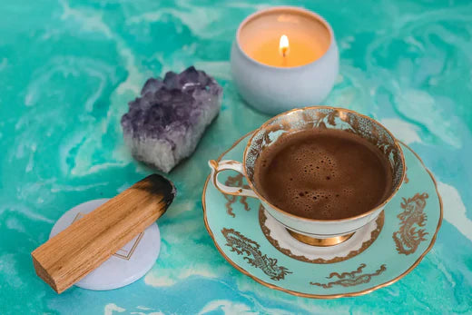Amethyst crystal cluster next to a cup of hot chocolate on an ornate turquoise cup and saucer, with a lit candle and a palo santo stick on a marble surface.