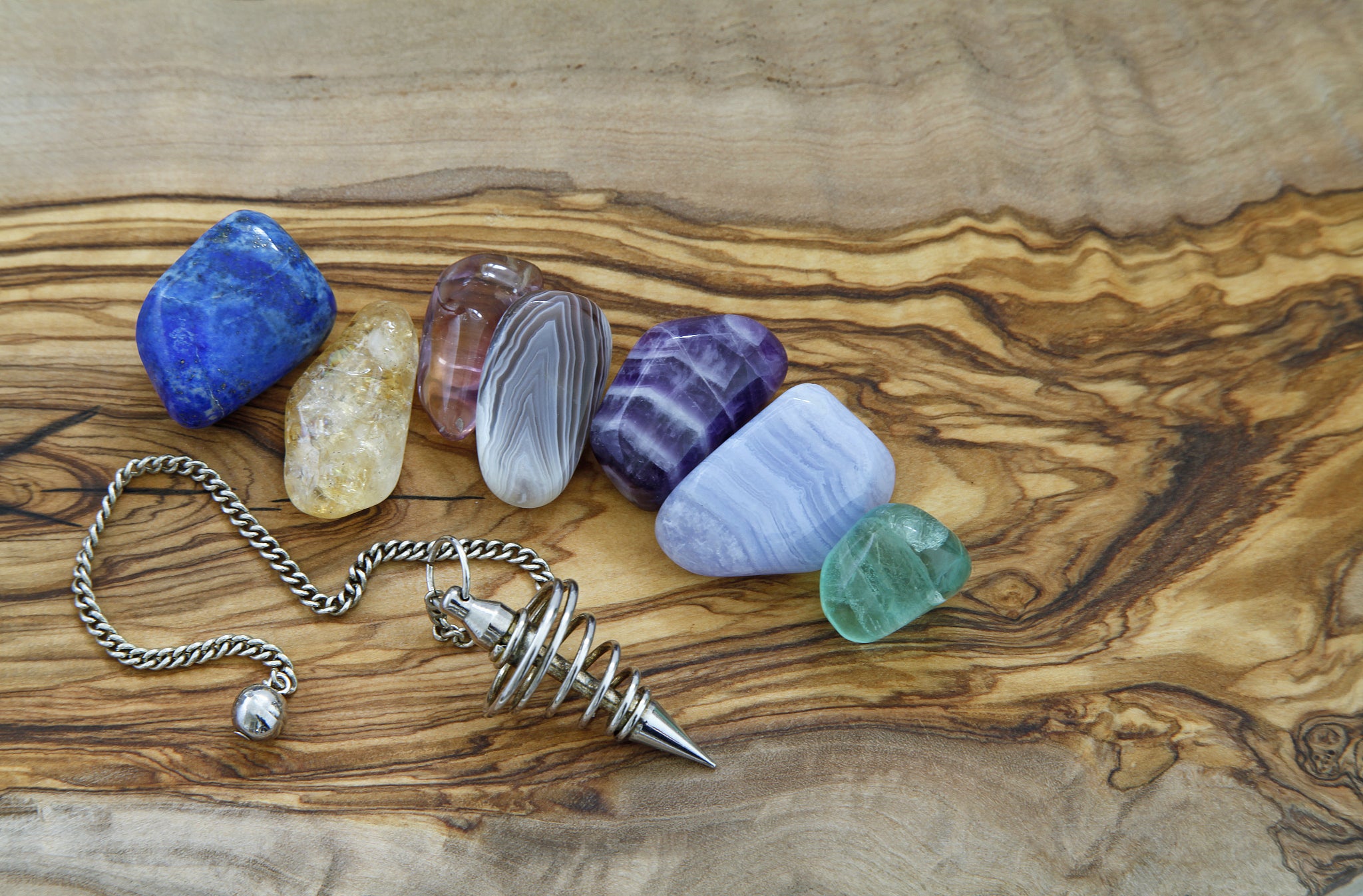 A variety of tumbled crystals, including lapis lazuli and amethyst, along with a metal pendulum on a wooden surface
